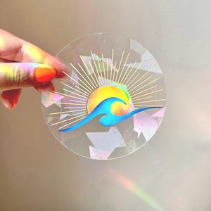 Peut inclure: Un autocollant transparent avec une finition holographique représentant un soleil jaune avec une vague bleue devant lui. Le soleil présente un dégradé de jaune, d'orange et de rouge. La vague est bleue avec une crête blanche. L'autocollant a un motif de rayons de soleil autour du soleil et de la vague.