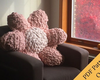 May include: A large pink and white crocheted flower pillow sits on a brown armchair. The pillow is made of chunky yarn and has a fluffy texture. The background is a window with fall foliage visible outside.