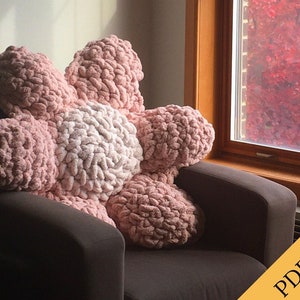 May include: A large pink and white crocheted flower pillow sits on a brown armchair. The pillow is made of chunky yarn and has a fluffy texture. The background is a window with fall foliage visible outside.