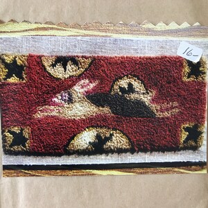 May include: A close-up of a red, white, and black rug with a whimsical design. The rug is made with a hooked technique and features a variety of shapes and patterns. The rug is labeled "Wylie Primitive Designs" and includes a contact number.