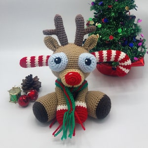 Peut inclure: Un jouet de renne en crochet portant une écharpe verte et rouge et des antennes en forme de canne à sucre. Le renne est assis devant un petit sapin de Noël.