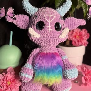 May include: A crocheted purple monster plush toy with rainbow fur, white horns, and large black eyes. The toy is sitting on a pink surface.