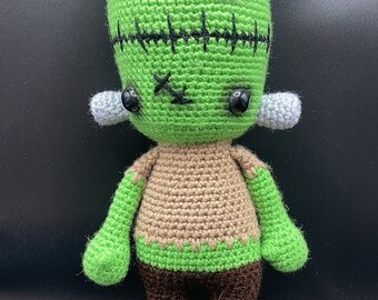 May include: Green crocheted Frankenstein monster doll with black stitches, brown trousers, and black eyes.