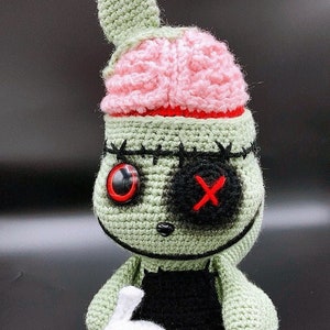 May include: A green crocheted plush toy with a pink brain hat, one red eye, and a black eye with a red X. The toy is holding a white crocheted acorn with a black skull face.