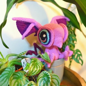 May include: A crocheted pink and purple creature with a large blue eye and a white pupil. The creature is sitting in a grey pot with green plants in the background.