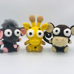 May include: Three crocheted stuffed animals: a gray elephant with pink ears, a yellow giraffe with brown spots, and a brown monkey with beige fur. The animals have large white eyes and are handmade.