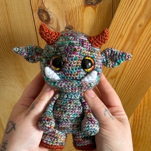 Op de afbeelding: Een gehaakt knuffelmonster met een groen, paars en oranje kleurenschema. Het monster heeft grote ogen, een witte mond en oranje hoorns.