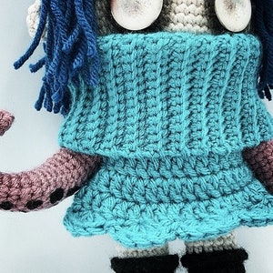 May include: A handmade crochet doll with a blue dress and blue yarn hair. The doll has large, round, white eyes and black boots. The doll's arm is a mauve color with black dots.