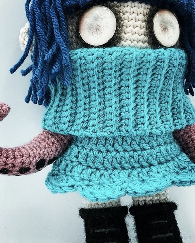 May include: A handmade crochet doll with a blue dress and blue yarn hair. The doll has large, round, white eyes and black boots. The doll's arm is a mauve color with black dots.