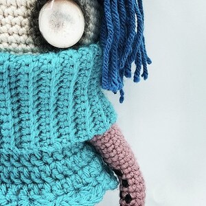 May include: Crocheted doll with a blue sweater, blue hair, and a large, round, clear eye. The doll's arm is pink with black accents. The doll is handmade and has a whimsical appearance.