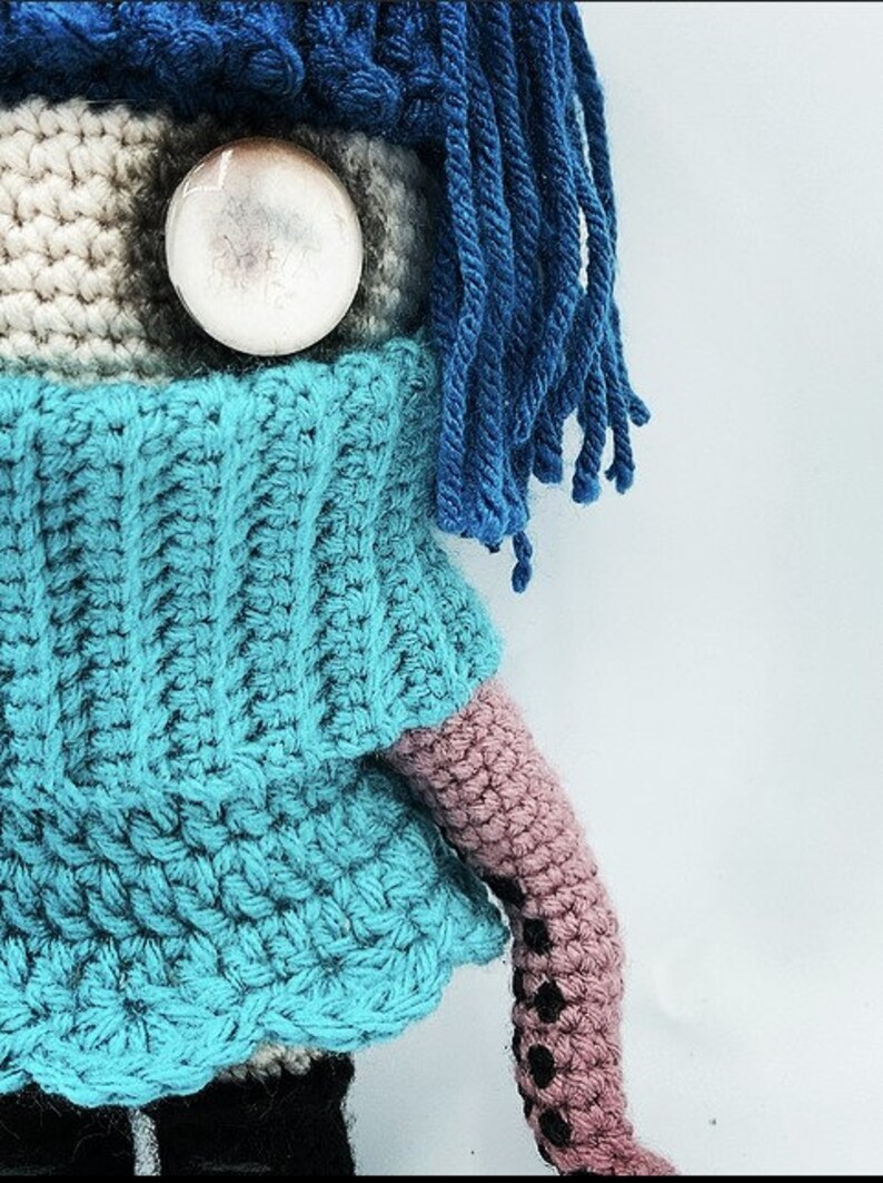 May include: Crocheted doll with a blue sweater, blue hair, and a large, round, clear eye. The doll's arm is pink with black accents. The doll is handmade and has a whimsical appearance.