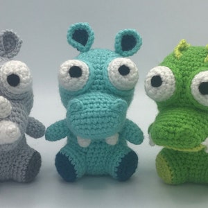 May include: Three crocheted stuffed animals: a gray rhinoceros, a teal hippopotamus, and a green crocodile. Each animal has large, white eyes with black pupils and a stitched smile.