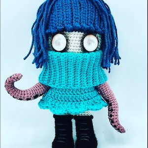 May include: A handmade crochet doll with blue hair and a matching turtleneck. The doll has large, round eyes and wears a turquoise dress. Its arms are pink with black spots, and it wears black boots. The doll is a unique, whimsical creation.