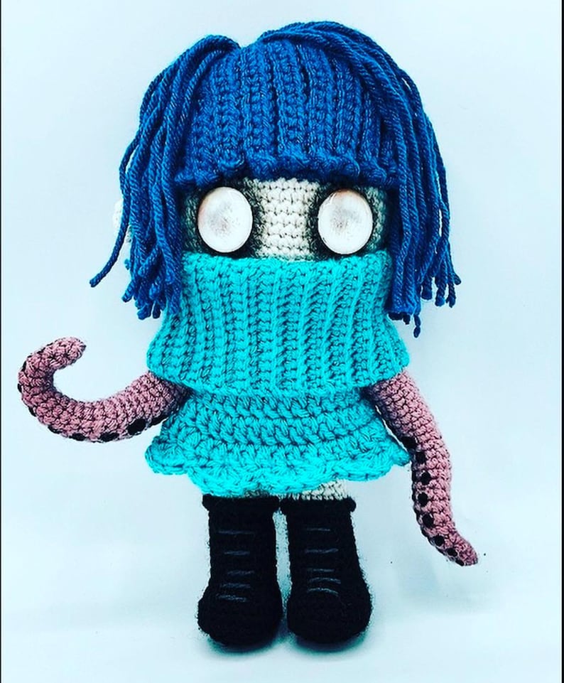 May include: A handmade crochet doll with blue hair and a matching turtleneck. The doll has large, round eyes and wears a turquoise dress. Its arms are pink with black spots, and it wears black boots. The doll is a unique, whimsical creation.