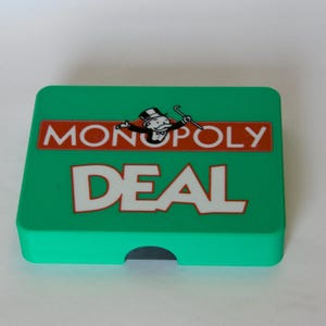 May include: A green Monopoly Deal card game box. The box features the Monopoly logo with the Monopoly man and the word "DEAL" in large white letters. The box is rectangular with a small cutout on one side.
