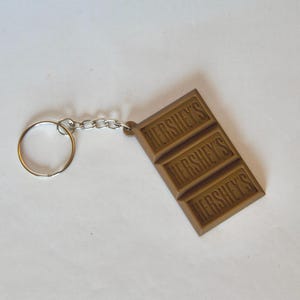 May include: A brown Hershey's chocolate bar keychain. The keychain features a rectangular chocolate bar design with the word "HERSHEY'S" embossed on each of the three sections. It includes a silver chain and a keyring.