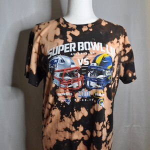 May include: A black and white bleached t-shirt with a graphic of two football helmets. The text on the shirt reads "Super Bowl LIII Atlanta, GA Patriots vs Rams 02.03.19".
