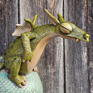 Könnte beinhalten: Eine detaillierte, grüne Keramikdrachenskulptur mit einem strukturierten, schuppigen Körper und großen, ausdrucksstarken Augen. Der Drache sitzt auf einer blau-grünen Kugel, wobei sein Kopf und seine Vorderbeine deutlich sichtbar sind. Der Hintergrund ist eine verwitterte Holzoberfläche.