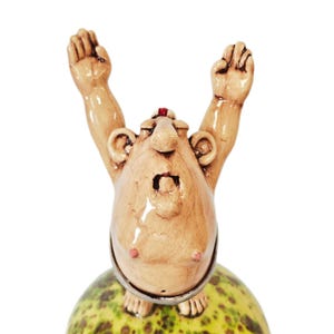 Könnte beinhalten: Eine skurrile Keramikfigur einer nackten Figur mit erhobenen Armen. Die Figur hat einen überraschten Gesichtsausdruck, mit einem kleinen roten Detail auf dem Kopf. Sie steht auf einem gesprenkelten grün-gelben Sockel mit einem silbernen Band um die Taille.