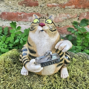 Könnte beinhalten: Eine Keramik-Katzenfigur mit beigefarbenem und schwarzem Streifenmuster, großen gelben Augen und einer rosa Nase. Die Katze hat einen überraschten Gesichtsausdruck und hält eine kleine, silberfarbene Metallsardinendose mit offenem Deckel in den Pfoten.