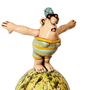May include: A whimsical ceramic figurine of a person in a striped swimsuit, arms outstretched. The figure wears sunglasses and a patterned head covering, standing atop a speckled, yellow-green sphere. The figurine has a surprised expression.