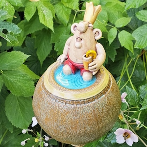 Könnte beinhalten: Eine skurrile Gartenfigur, die eine Cartoonfigur mit Krone und roter Hose zeigt, die in einem blauen Pool entspannt. Die Figur hält ein Getränk mit einer Zitronenscheibe. Die Statue befindet sich in einem strukturierten, runden, braunen Topf.