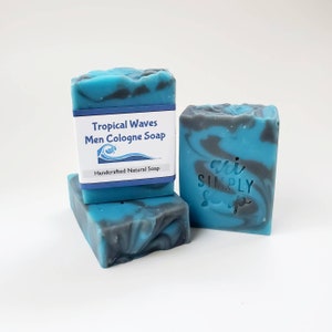 May include: Three bars of handcrafted soap with a tropical wave design. The soap is blue with gray marbling and a white label that reads "Tropical Waves Men Cologne Soap". The soap is a natural product.