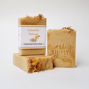 May include: Three bars of handmade soap, one with a label that reads "Calendula Oatmeal Handcrafted Natural Soap". The soap is a light beige color and has a natural, earthy look. The other two bars have the words "ari SIMPLY Soap" stamped on them.