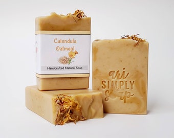 May include: Three bars of handmade soap, one with a label that reads "Calendula Oatmeal Handcrafted Natural Soap". The soap is a light beige color and has a natural, earthy look. The other two bars have the words "ari SIMPLY Soap" stamped on them.