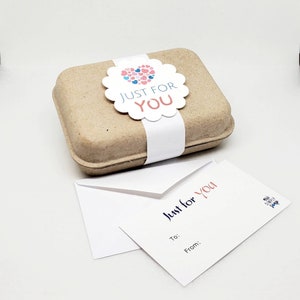 May include: A small brown cardboard box with a white label that says "Just for You" and a heart-shaped sticker with pink and blue hearts. The box is sitting on a white surface next to a white card with the text "Just for You" and a small logo that says "My Sweet Shop".