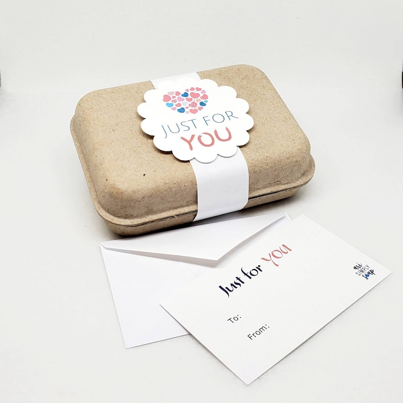May include: A small brown cardboard box with a white label that says "Just for You" and a heart-shaped sticker with pink and blue hearts. The box is sitting on a white surface next to a white card with the text "Just for You" and a small logo that says "My Sweet Shop".