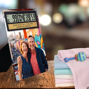 May include: A digital countdown clock with the text "Adam & Eve will turn into Grandpa & Grandma" sits above a photo of a smiling couple. A stack of pastel baby blankets and a rattle are on the right.