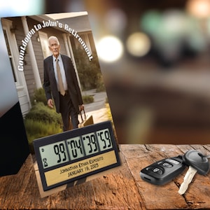 May include: A black countdown clock with a digital display showing "99 '04 '39 '59". The clock has a gold plaque with the text "JOHNATHAN ETHAN ESPOSITO JANUARY 19, 2025". The clock is sitting on a wooden surface with a photo of a man in a suit standing in front of a house. The photo has the text "Countdown to John's Retirement" around the edge.