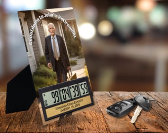 May include: A black countdown clock with a digital display showing "99 '04 '39 '59". The clock has a gold plaque with the text "JOHNATHAN ETHAN ESPOSITO JANUARY 19, 2025". The clock is sitting on a wooden surface with a photo of a man in a suit standing in front of a house. The photo has the text "Countdown to John's Retirement" around the edge.