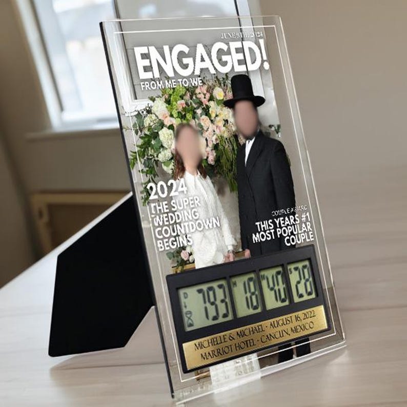 May include: A clear acrylic countdown clock with a black digital display showing the numbers 793, 18, 40, and 28. The clock is decorated with a photo of a couple in front of a floral backdrop. The text "ENGAGED!" is printed in white on the top of the clock. The text "FROM ME TO WE" is printed in white below the word "ENGAGED!" The text "2024" is printed in white below the photo. The text "THE SUPER WEDDING COUNTDOWN BEGINS" is printed in white below "2024". The text "COUPLE AFASC" is printed in white on the right side of the clock. The text "THIS YEARS #1 MOST POPULAR COUPLE" is printed in white below "COUPLE AFASC". The text "MICHELLE & MICHAEL - AUGUST 16, 2022 MARRIOT HOTEL - CANCUN, MEXICO" is printed in gold on the bottom of the clock.
