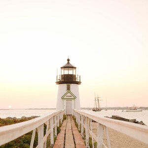 May include: A white lighthouse with a black top and a green door frame stands at the end of a wooden pier. The pier has white railings. Boats are in the water in the background. The sky is a soft peach color.