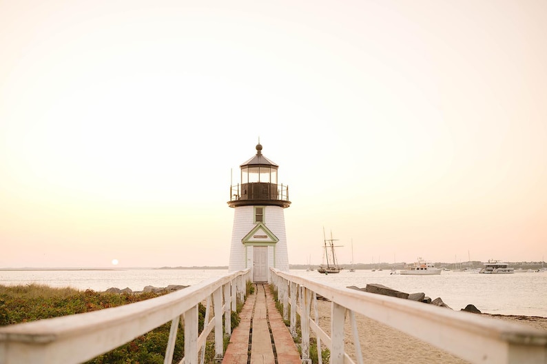 May include: A white lighthouse with a black top and a green door frame stands at the end of a wooden pier. The pier has white railings. Boats are in the water in the background. The sky is a soft peach color.