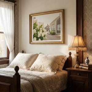 May include: A bedroom scene featuring a wooden bed with cream-colored bedding and embroidered pillows. A framed landscape painting hangs above the bed. A lamp with a fringed shade sits on a wooden nightstand, next to a stack of books and a small vase.