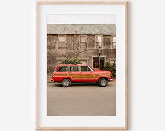 May include: A framed print featuring a vintage red and wood-paneled SUV parked on a street in front of a weathered wooden building. The vehicle has a green bundle on its roof. The scene is captured in natural light.