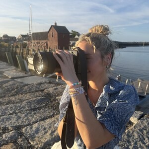 May include: A black digital camera with a wide lens and a dark leather strap, held by a person wearing a blue floral print blouse with ruffled sleeves and white trousers, adorned with several colourful beaded bracelets on the wrist.