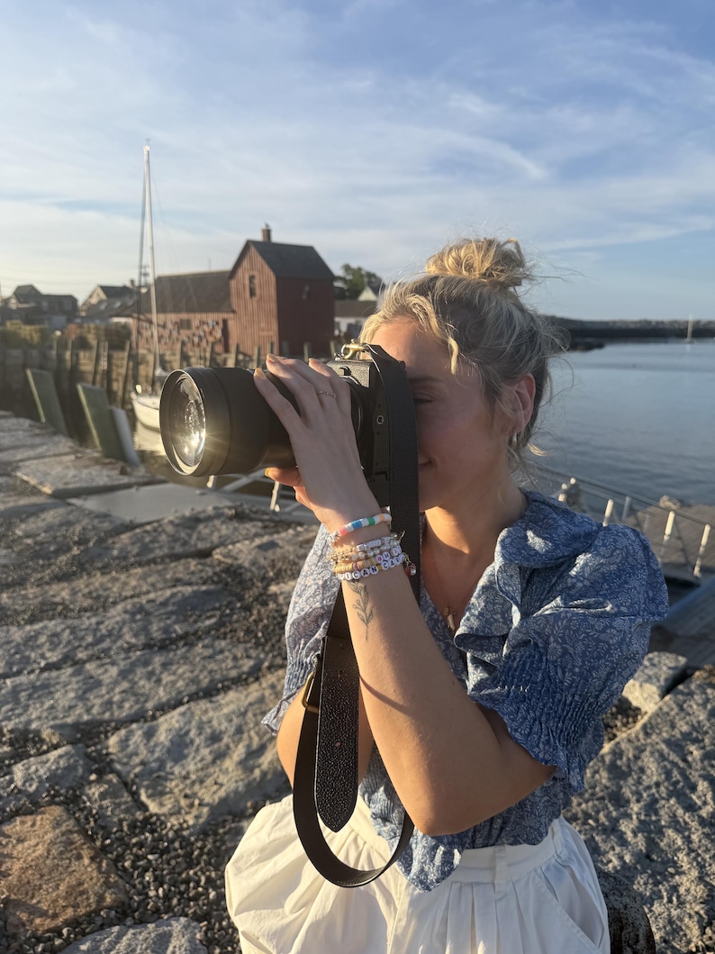 May include: A black digital camera with a wide lens and a dark leather strap, held by a person wearing a blue floral print blouse with ruffled sleeves and white trousers, adorned with several colourful beaded bracelets on the wrist.