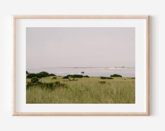 May include: A framed landscape print depicting a coastal scene. The image shows a field of tall green grass in the foreground, with a body of water and a distant shoreline. A boat is visible on the water under a cloudy sky.