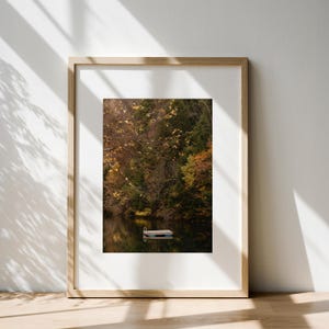 May include: A framed vertical wall art print featuring a serene lake scene with a small floating dock centred on dark water, surrounded by a dense forest displaying autumn foliage in shades of deep green, golden yellow, and burnt orange.