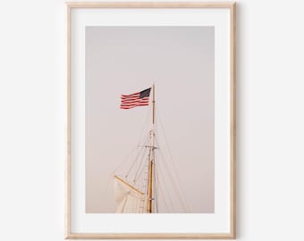 May include: A framed print featuring a sailboat mast with the American flag waving in the wind. The flag displays red and white stripes with a blue canton and white stars. The mast is made of wood and has rigging.