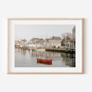 May include: A red rowboat with the word "MANUOCHET" written on the side, floating in a calm harbor with a blurred background of houses and docks.