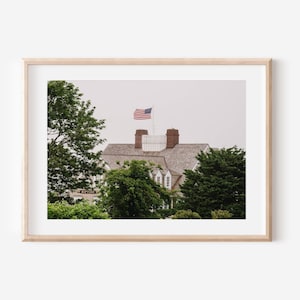 May include: A framed photograph of a building with a gray roof and brick chimneys. An American flag flies from a pole on the roof. Green trees and foliage surround the building, and the sky is overcast.