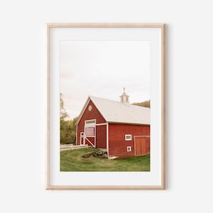 Puede incluir: Impresión enmarcada de un granero rojo con techo blanco y bandera estadounidense. El granero se encuentra frente a hierba verde y árboles, con un cielo nublado. La impresión está en un marco de madera clara.