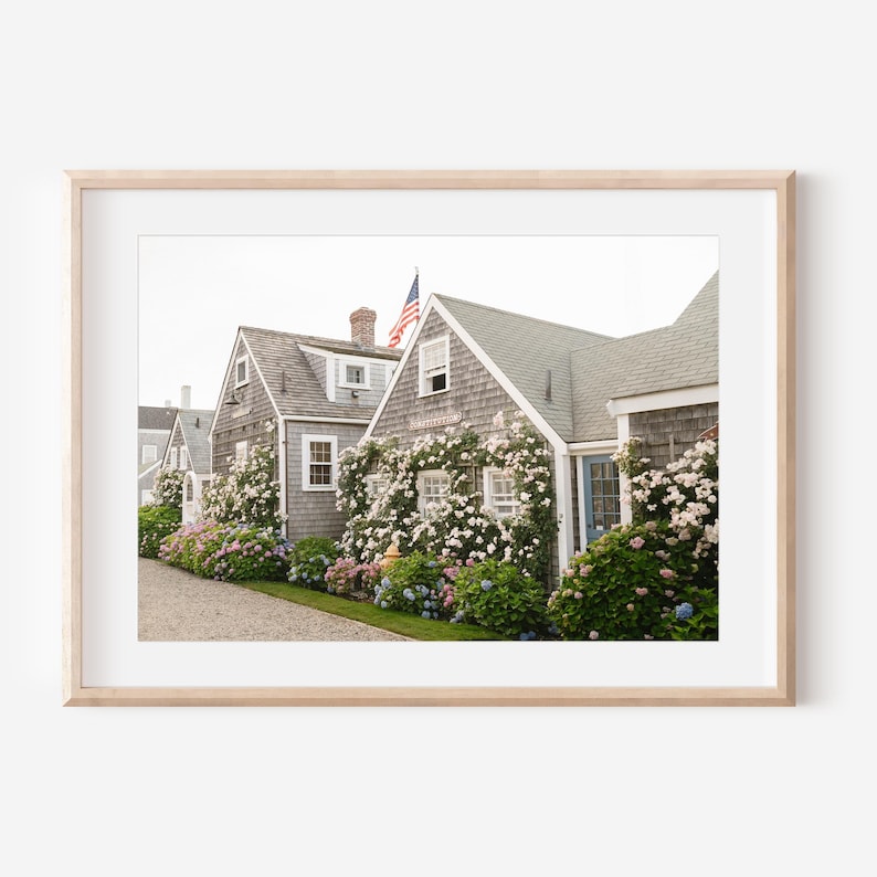 May include: A framed print featuring a row of charming gray-shingled houses with white trim. The houses are adorned with climbing white roses and colorful hydrangeas. An American flag waves in the background, adding a patriotic touch to the coastal scene.