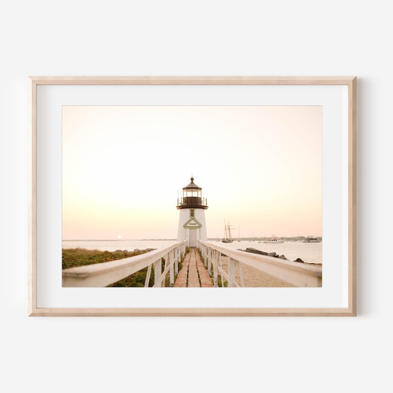 May include: A framed print featuring a lighthouse at sunset. The white lighthouse with a black top is at the end of a wooden walkway with white railings. The sky is a soft yellow and pink, and the water is calm.
