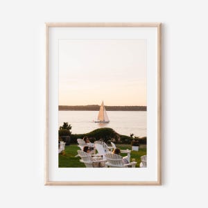May include: A framed photograph of a serene coastal scene. A sailboat glides across the water under a soft, golden sunset. White Adirondack chairs are arranged on a green lawn, with people enjoying the view.